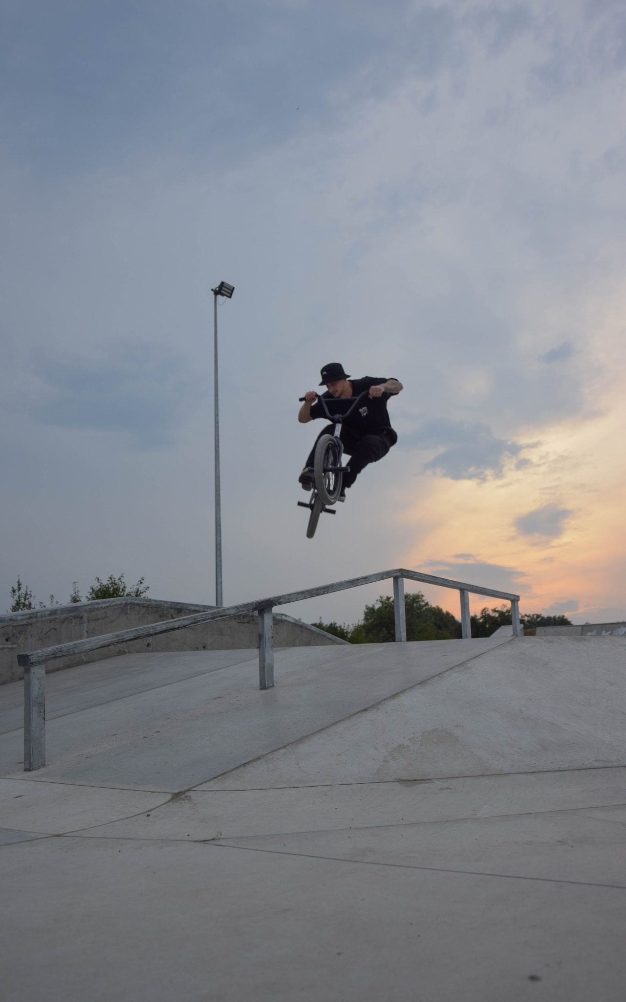 Mężczyzna w czarnym ubraniu i kapeluszu wykonuje trik na rowerze BMX w skateparku, unosząc się nad metalową barierką na tle zachodzącego słońca.