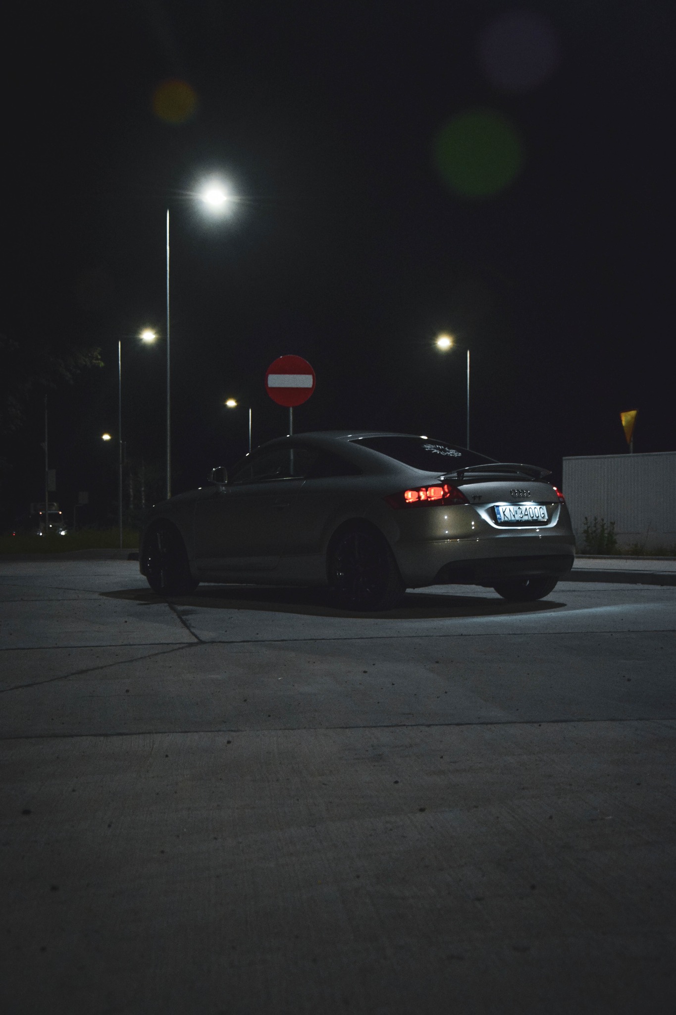 Audi TT w kolorze szampańskim, zaparkowane nocą na parkingu pod lampami ulicznymi, widok z tyłu, na tylnej szybie naklejki, widoczny znak zakazu wjazdu.