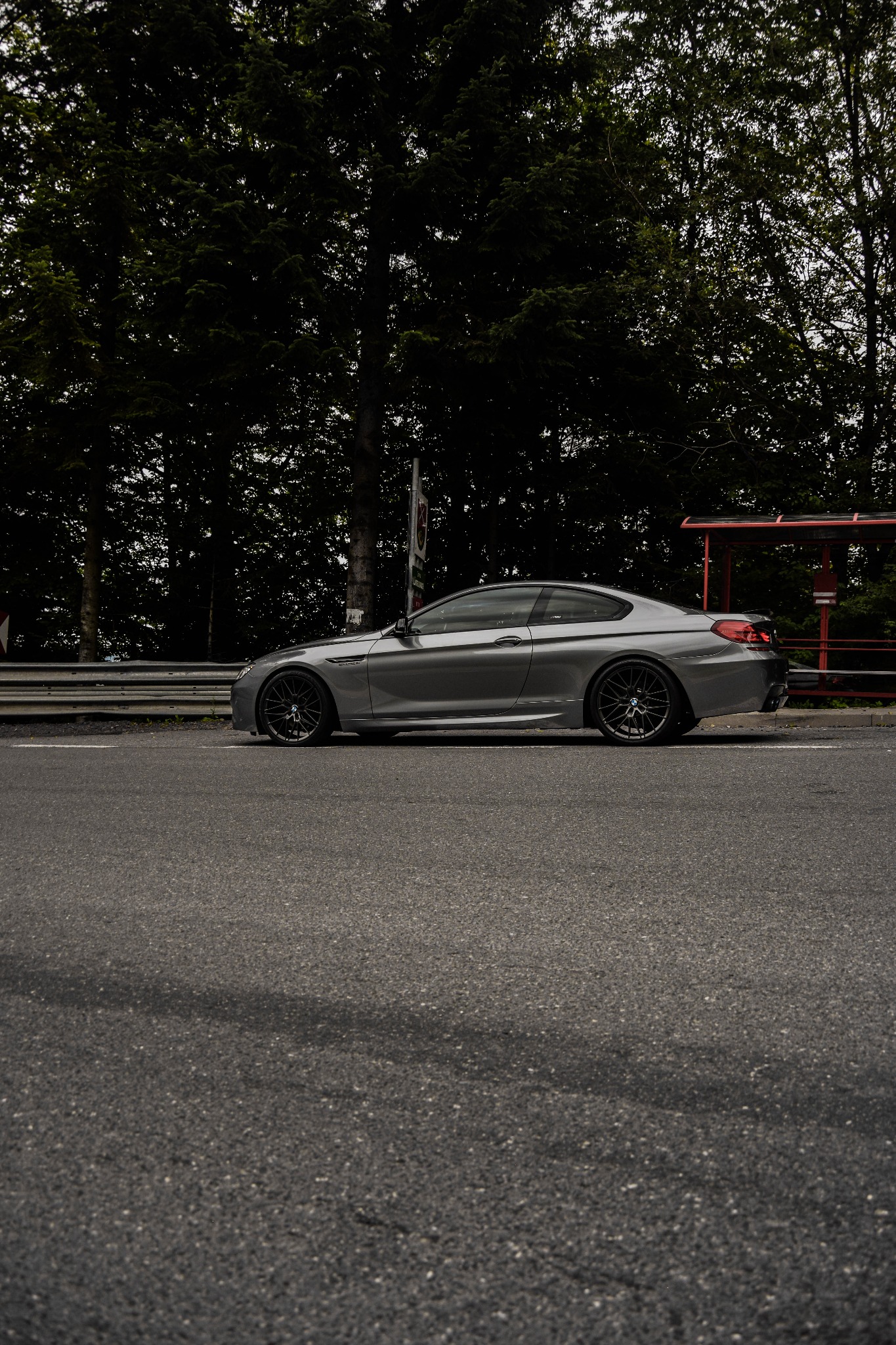 Szary samochód sportowy BMW serii 6 Coupe zaparkowany na poboczu drogi z ciemnymi felgami, na tle drzew i przystanku autobusowego.
