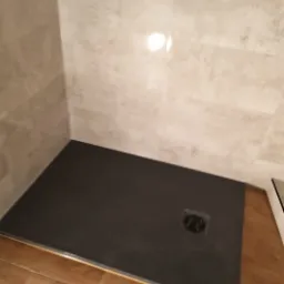 Czarny brodzik z odpływem liniowym ułożony w rogu łazienki z beżowymi płytkami imitującymi kamień i drewnopodobną podłogą.