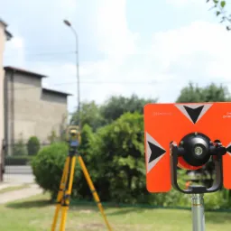 Pomarańczowy cel geodezyjny z czarnymi trójkątami i napisem '0 mm OFFSET' na tle zielonej trawy, drzew i budynku. W tle widoczny żółty teodolit na statywie.