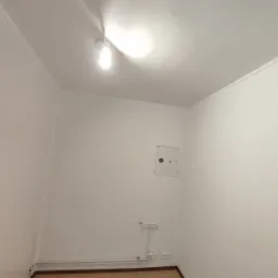 Pusta, biała przestrzeń z zamontowaną żarówką, widoczna skrzynka elektryczna i rurki przy podłodze.