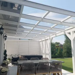 Biała konstrukcja z przeszklonym dachem nad tarasem, stół z plecionymi krzesłami, oświetlenie zewnętrzne w stylu retro, zadbany trawnik w tle.