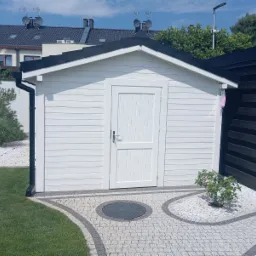 Biała, drewniana konstrukcja z czarnym dachem, przypominająca domek narzędziowy, otoczona trawnikiem i dekoracyjnym kamieniem. Widoczny fragment brukowanej ścieżki z ciemniejszym elementem centralnym.