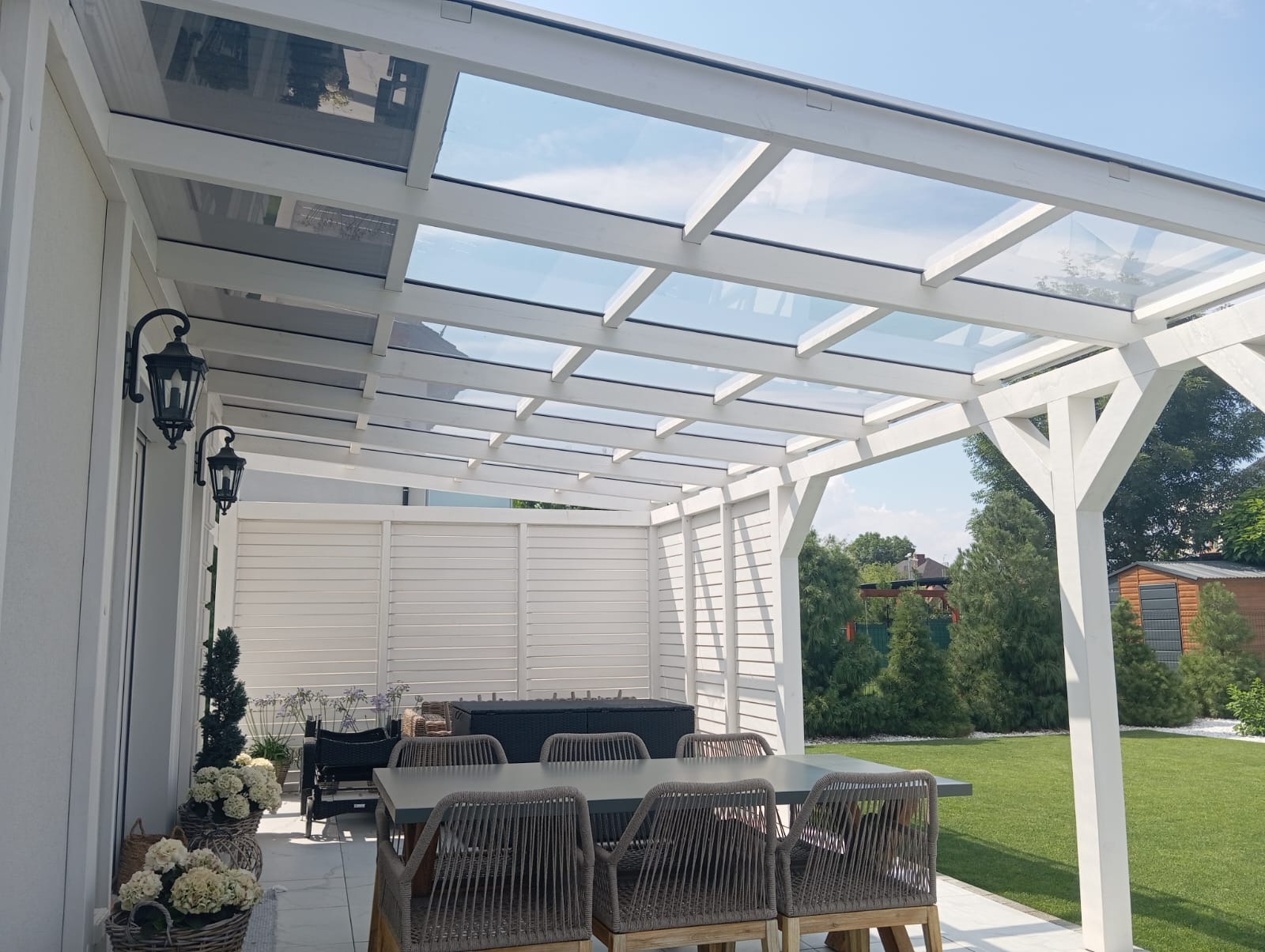 Biała konstrukcja z przeszklonym dachem nad tarasem, stół z plecionymi krzesłami, oświetlenie zewnętrzne w stylu retro, zadbany trawnik w tle.