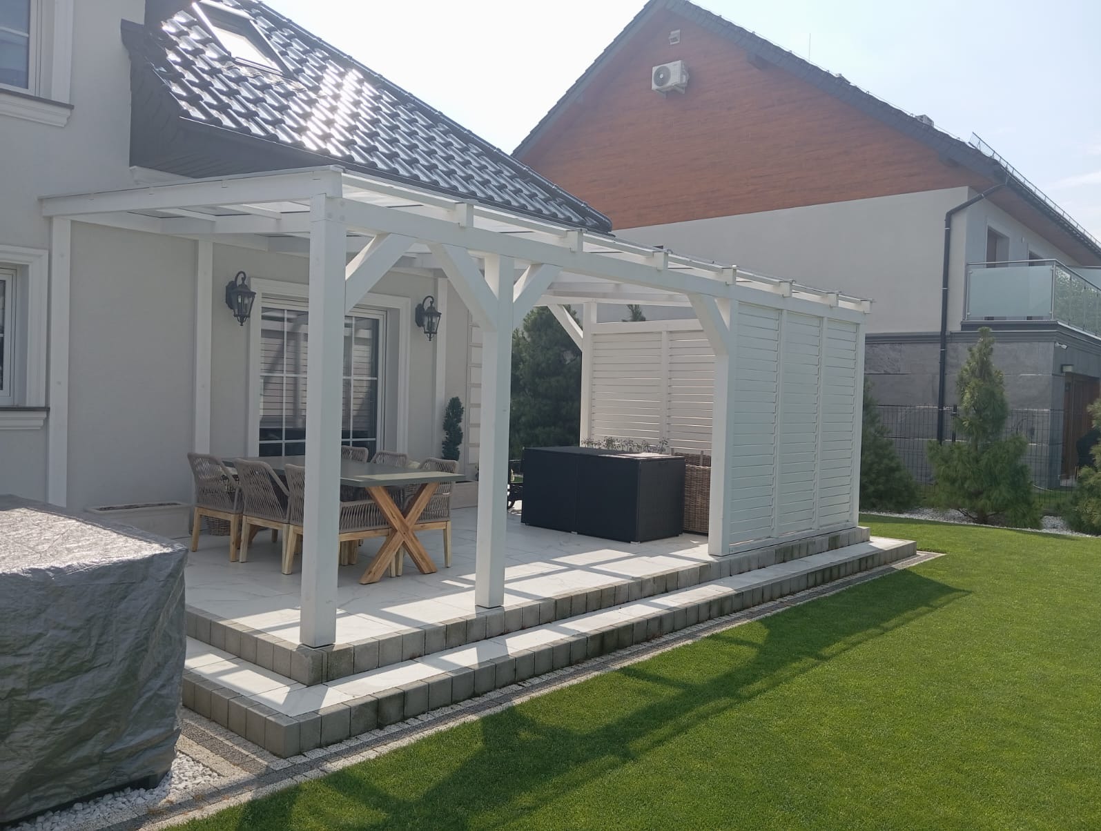 Biała konstrukcja z drewna z zadaszeniem, częściowo osłonięta ażurowymi panelami, na podwyższonym tarasie z jasnych płyt, zestaw mebli ogrodowych ze stołem z drewnianym krzyżowym stelażem, widok...