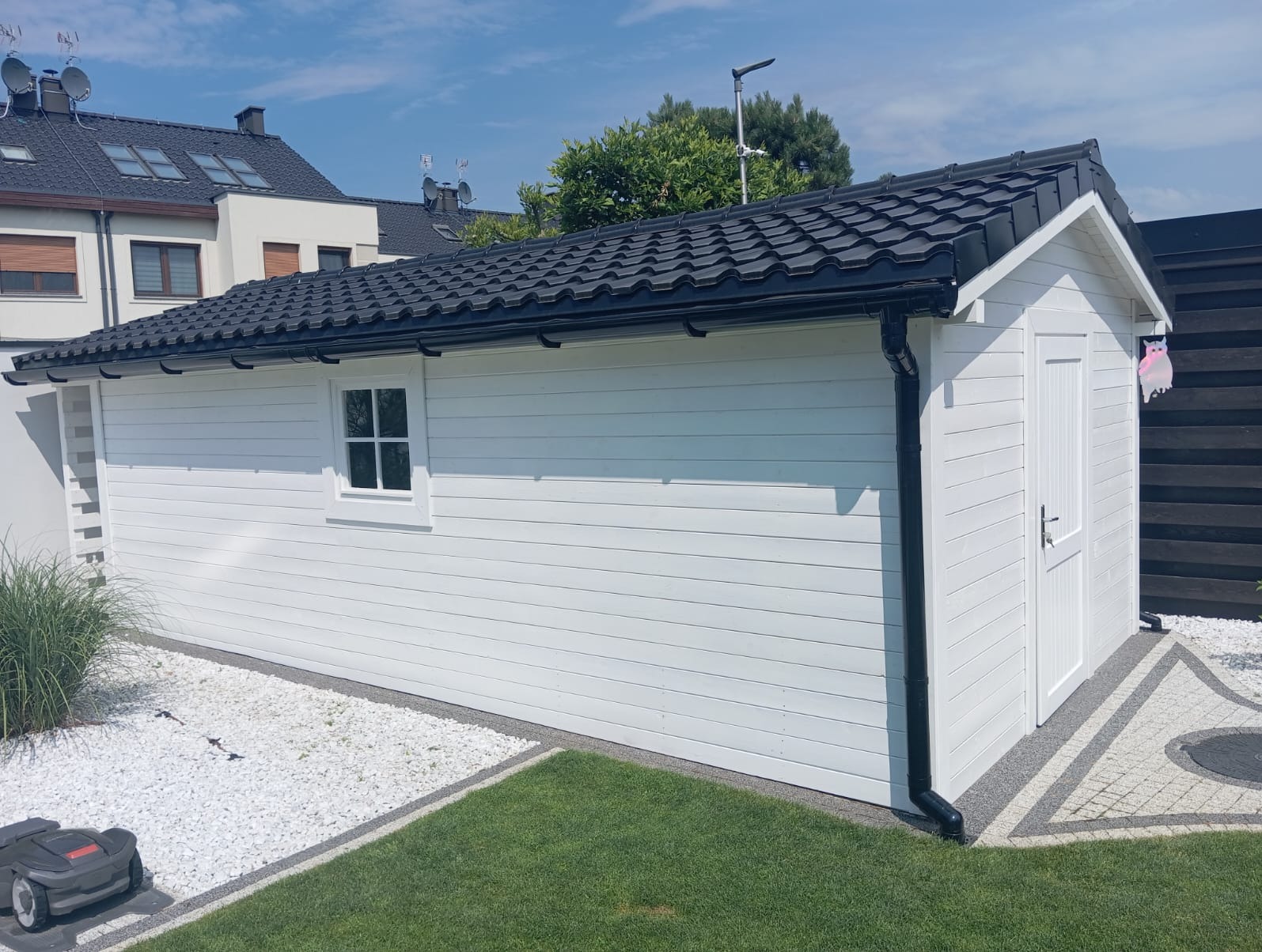 Biały drewniany domek narzędziowy z czarnym dachem na tle zielonego trawnika i dekoracyjnego żwiru, z widocznym robotem koszącym i ozdobą w kształcie sowy na drzwiach.