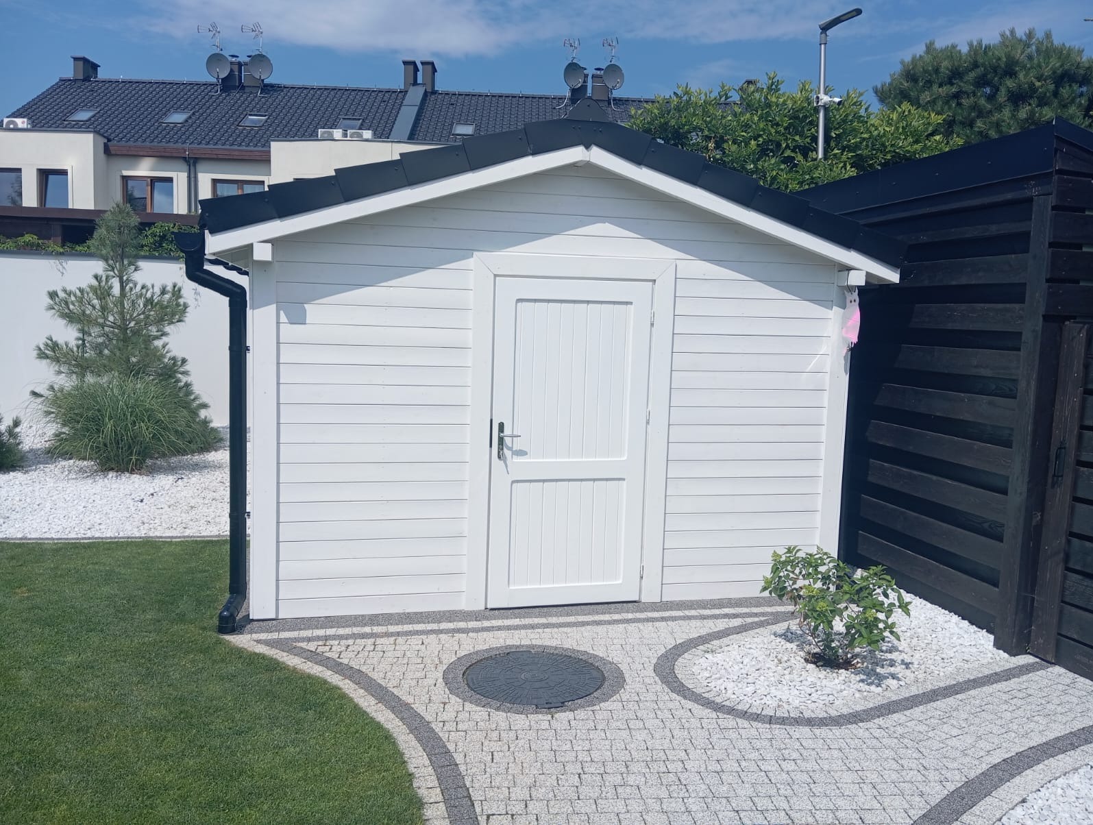 Biała, drewniana konstrukcja z czarnym dachem, przypominająca domek narzędziowy, otoczona trawnikiem i dekoracyjnym kamieniem. Widoczny fragment brukowanej ścieżki z ciemniejszym elementem centralnym.