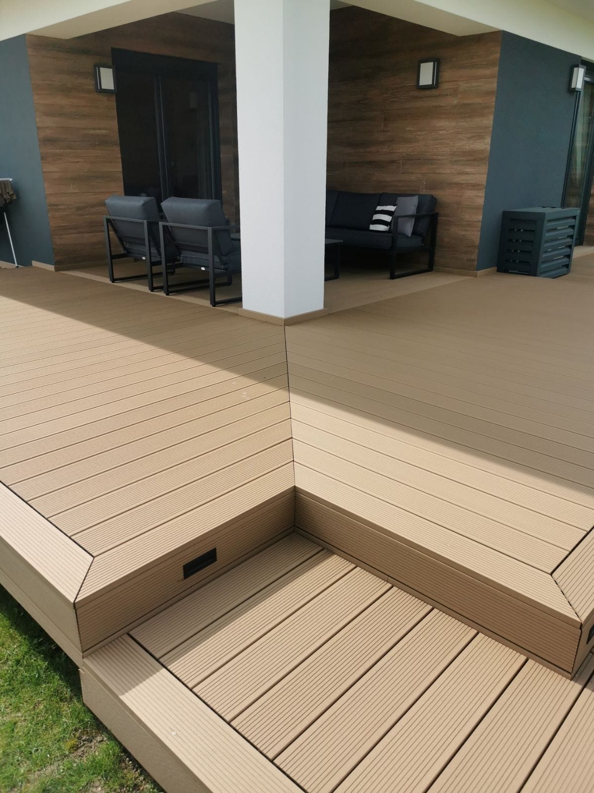 Beżowy taras z desek kompozytowych z niskim stopniem, oświetleniem punktowym w stopniu i nowoczesnymi meblami wypoczynkowymi w tle.