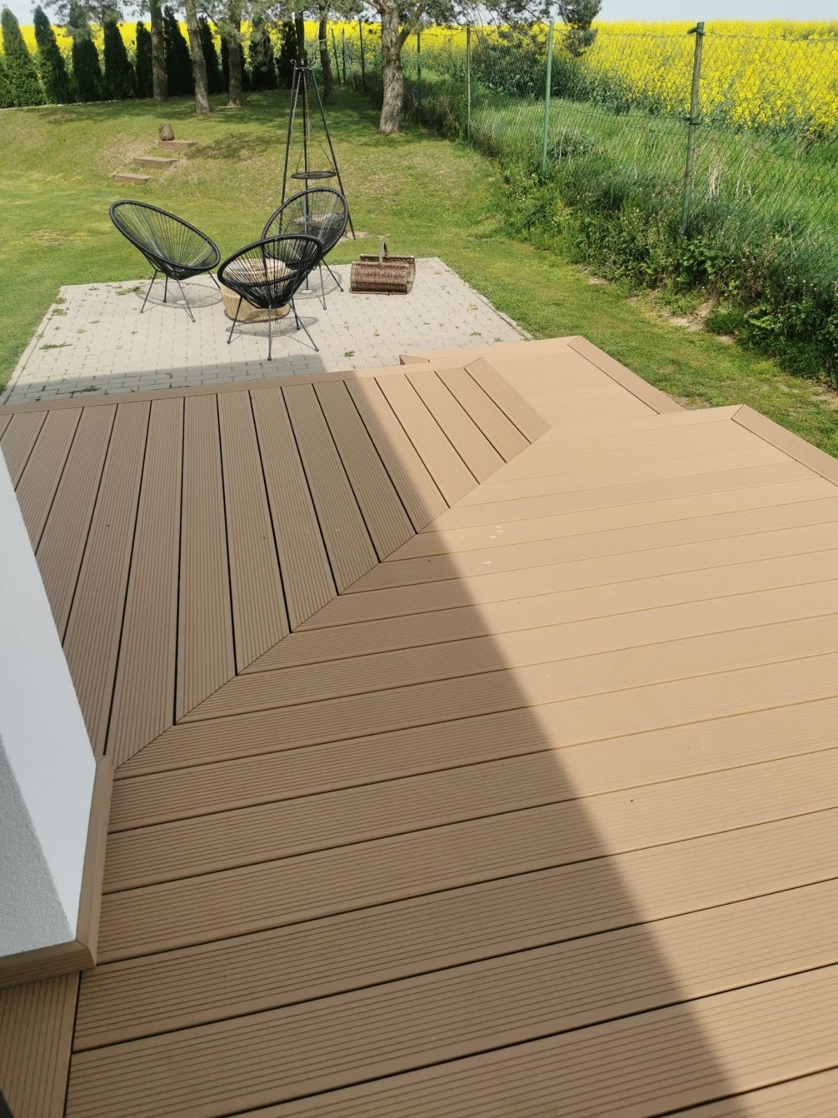 Beżowy taras z desek kompozytowych prowadzący do betonowego patio z trzema czarnymi krzesłami typu Acapulco i koszem piknikowym na tle kwitnącego pola rzepaku.