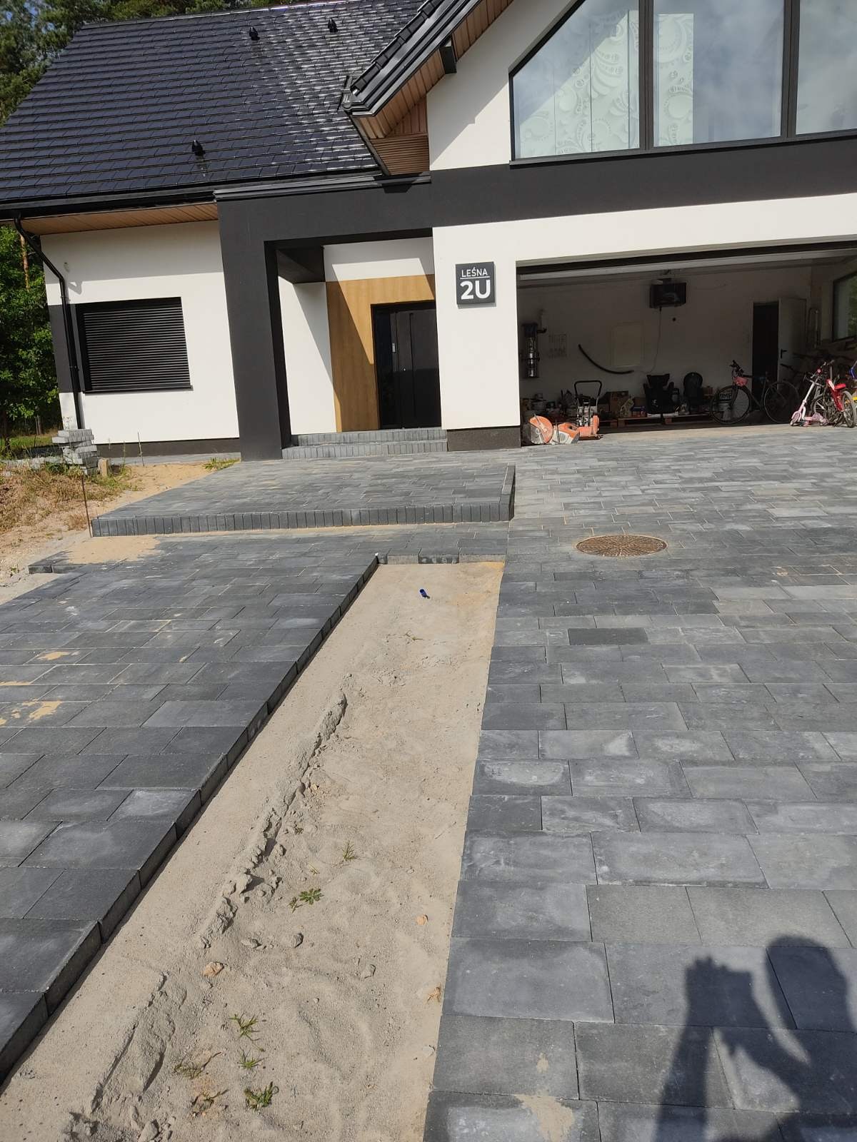 Częściowo ułożona, szara kostka brukowa przed nowoczesnym domem z garażem; widoczna piaskowa podsypka i narzędzia w garażu.