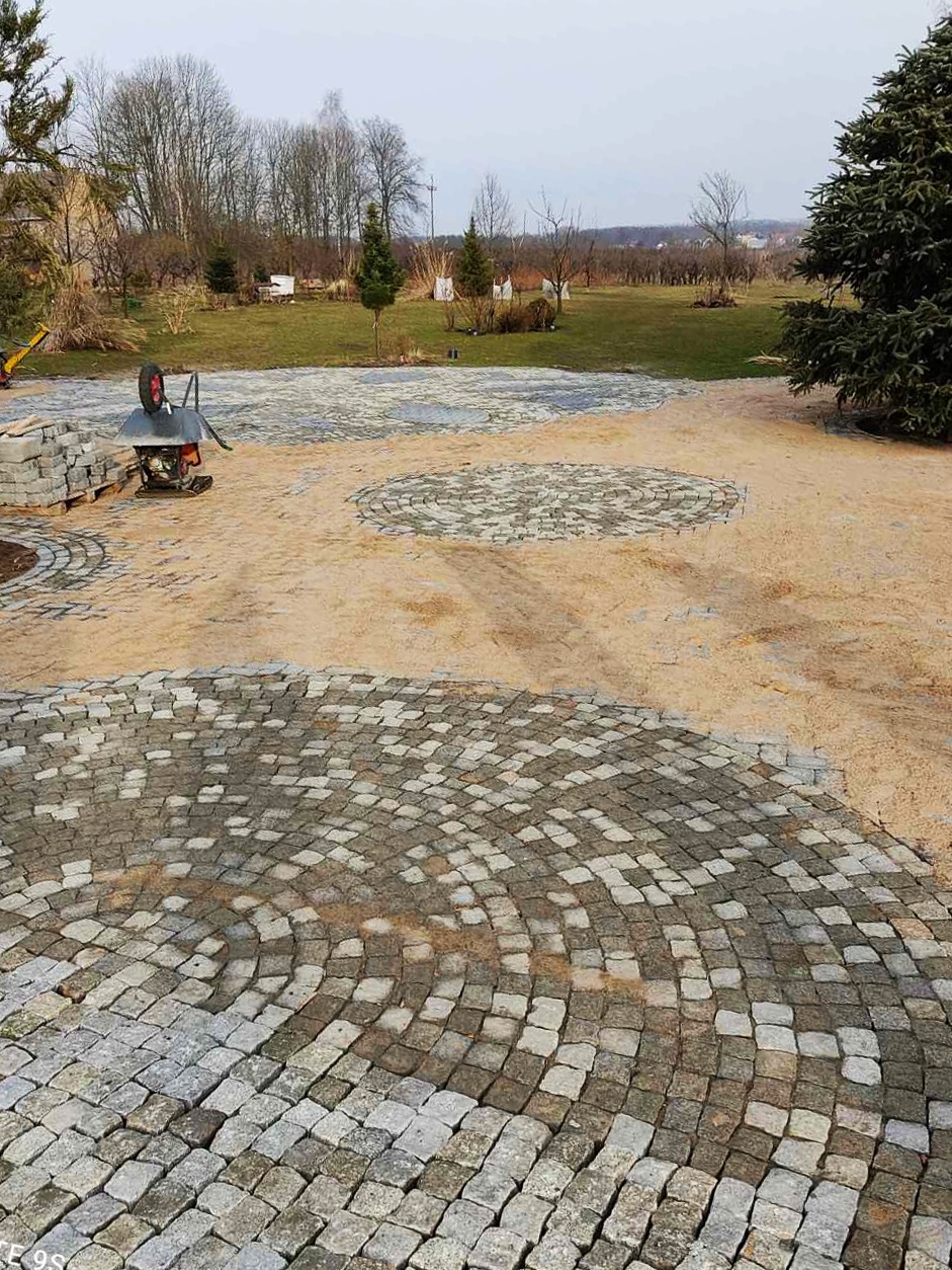 Układanie okręgów z kostki granitowej na piaszczystym podłożu, widoczna taczka z ładunkiem kostki i paleta z materiałem w tle, etap prac na posesji.