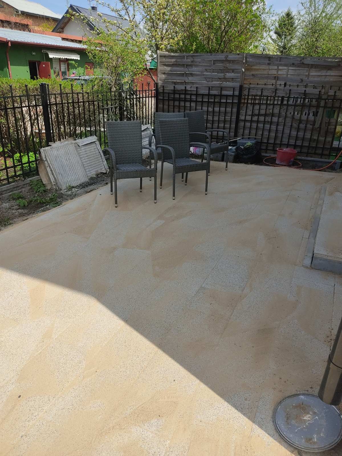 Nowa, jasna nawierzchnia tarasu z kostki z czterema szarymi krzesłami ogrodowymi, widoczne przygotowane płyty kostki, czarne ogrodzenie i elementy wyposażenia ogrodowego w tle.