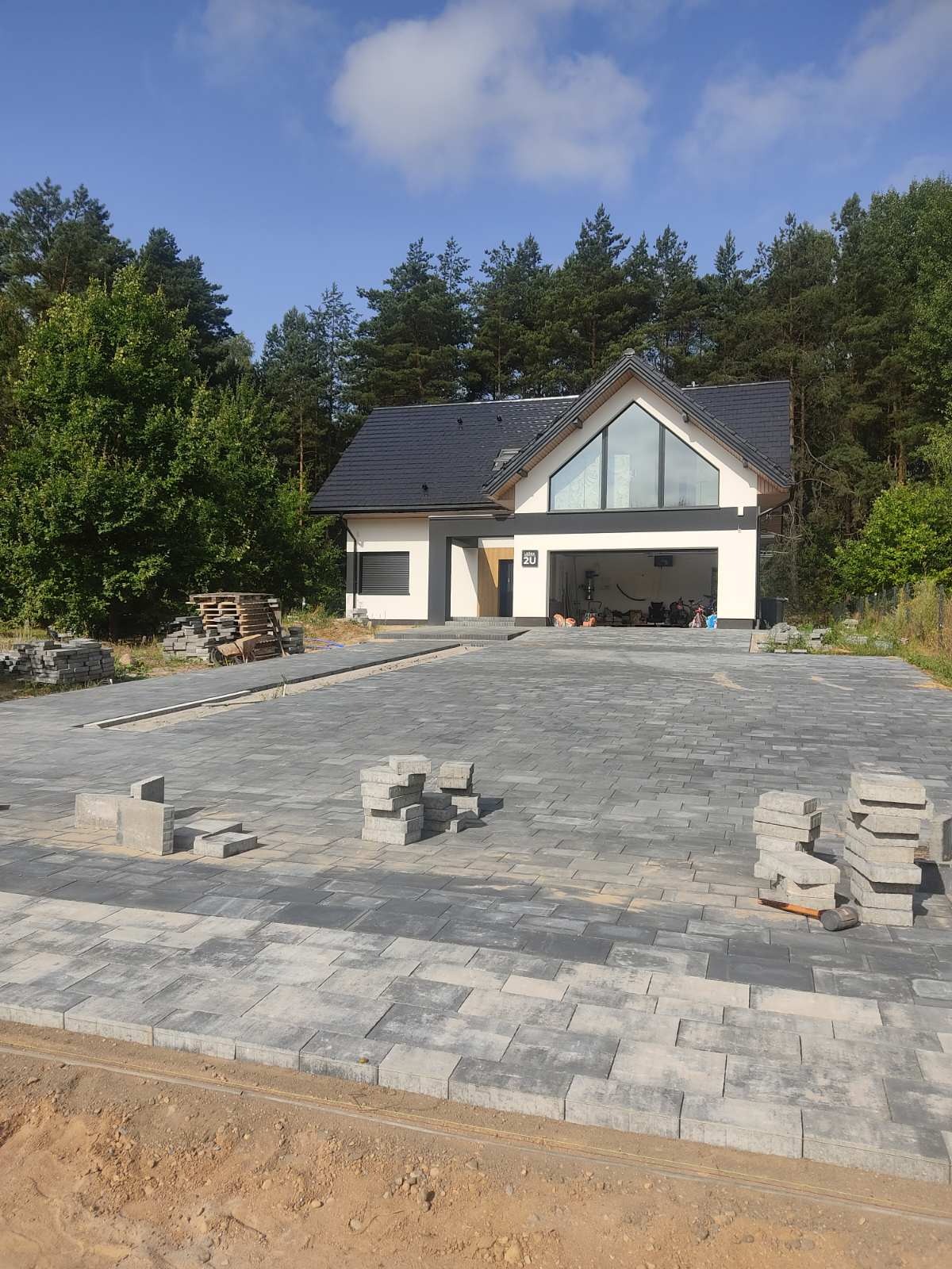 Kładzenie szarej kostki brukowej na podjeździe przed nowoczesnym domem z dużym przeszkleniem, widoczne stosy kostki i młotek.