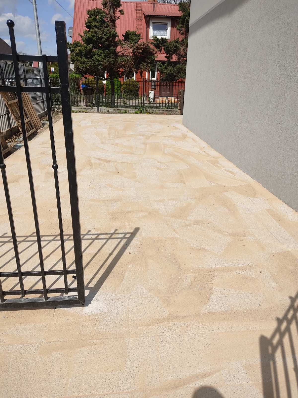 Jasna kostka granitowa ułożona na podjeździe, widoczna brama wjazdowa z czarnymi prętami, tło z domem z czerwoną dachówką.