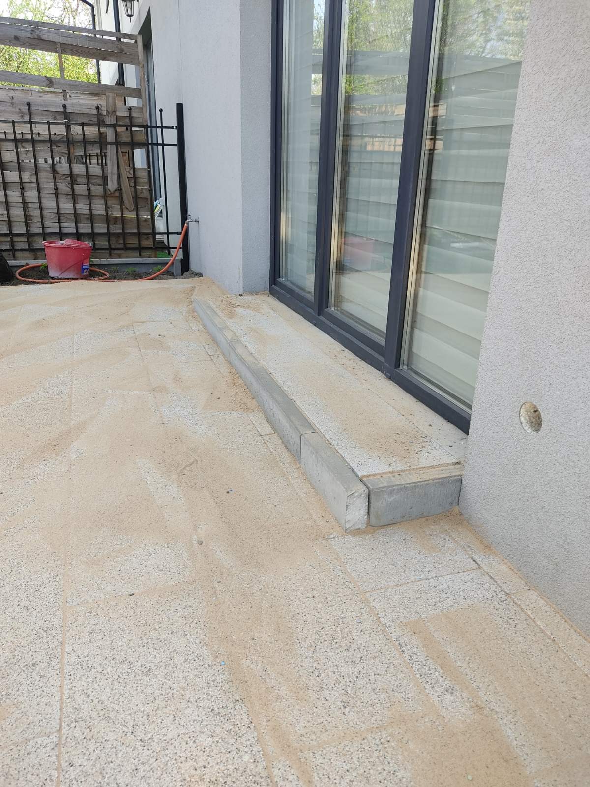 Granitowa kostka brukowa w trakcie układania, zasypana piaskiem przy nowoczesnym domu z dużymi oknami, widoczny fragment obrzeża i czerwone wiadro w tle.