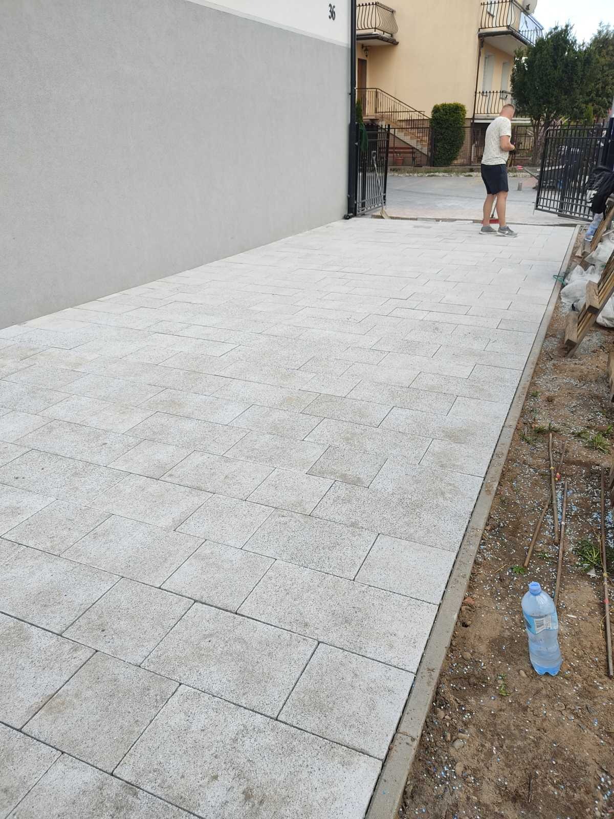 Świeżo ułożona, jasnoszara kostka granitowa na podjeździe, widoczna butelka wody i narzędzia wzdłuż krawędzi, osoba w tle.