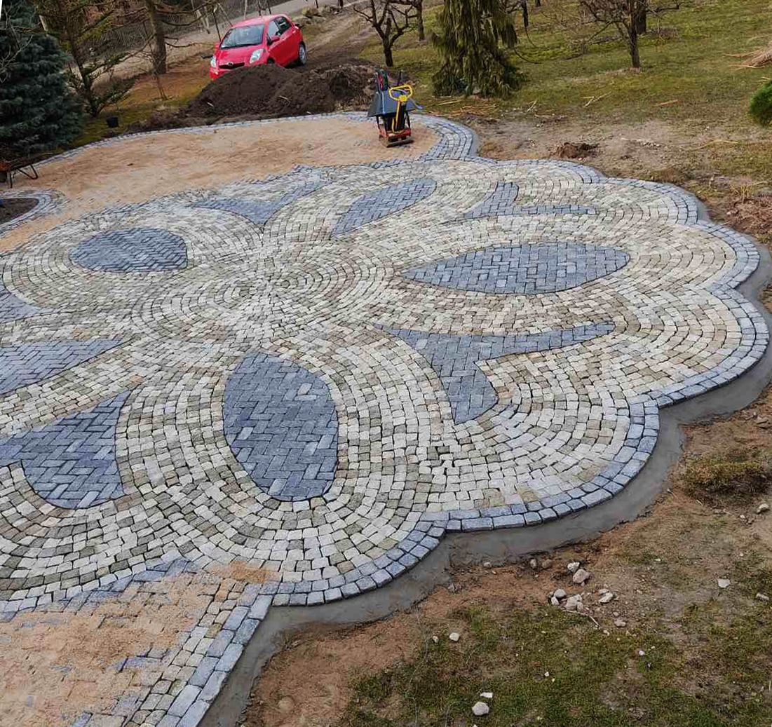 Układanie wzoru kwiatowego z kostki granitowej w ogrodzie, widoczna zagęszczarka w tle, czerwony samochód na wzgórzu