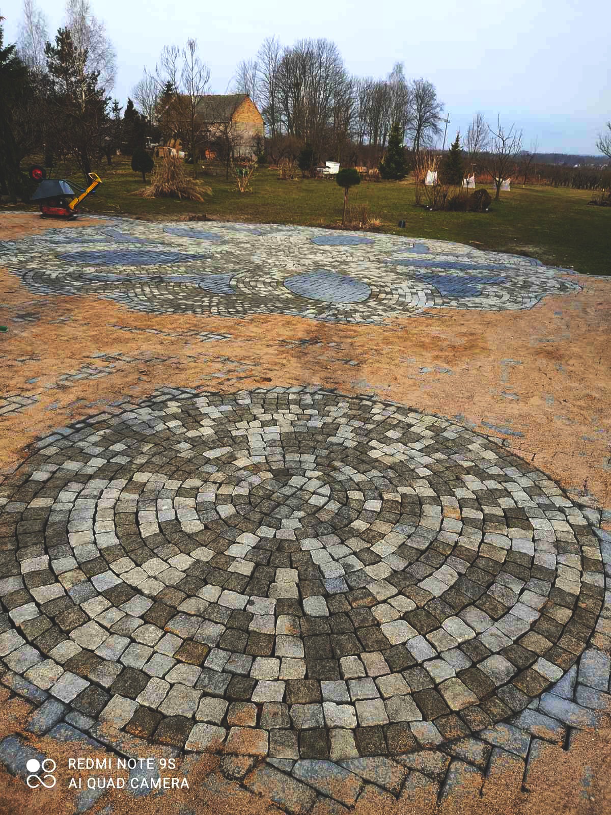 Okrąg z kostki granitowej w odcieniach szarości ułożony na piasku, w tle trawnik z drzewami i budynkiem gospodarczym, widoczna maszyna do ubijania ziemi.