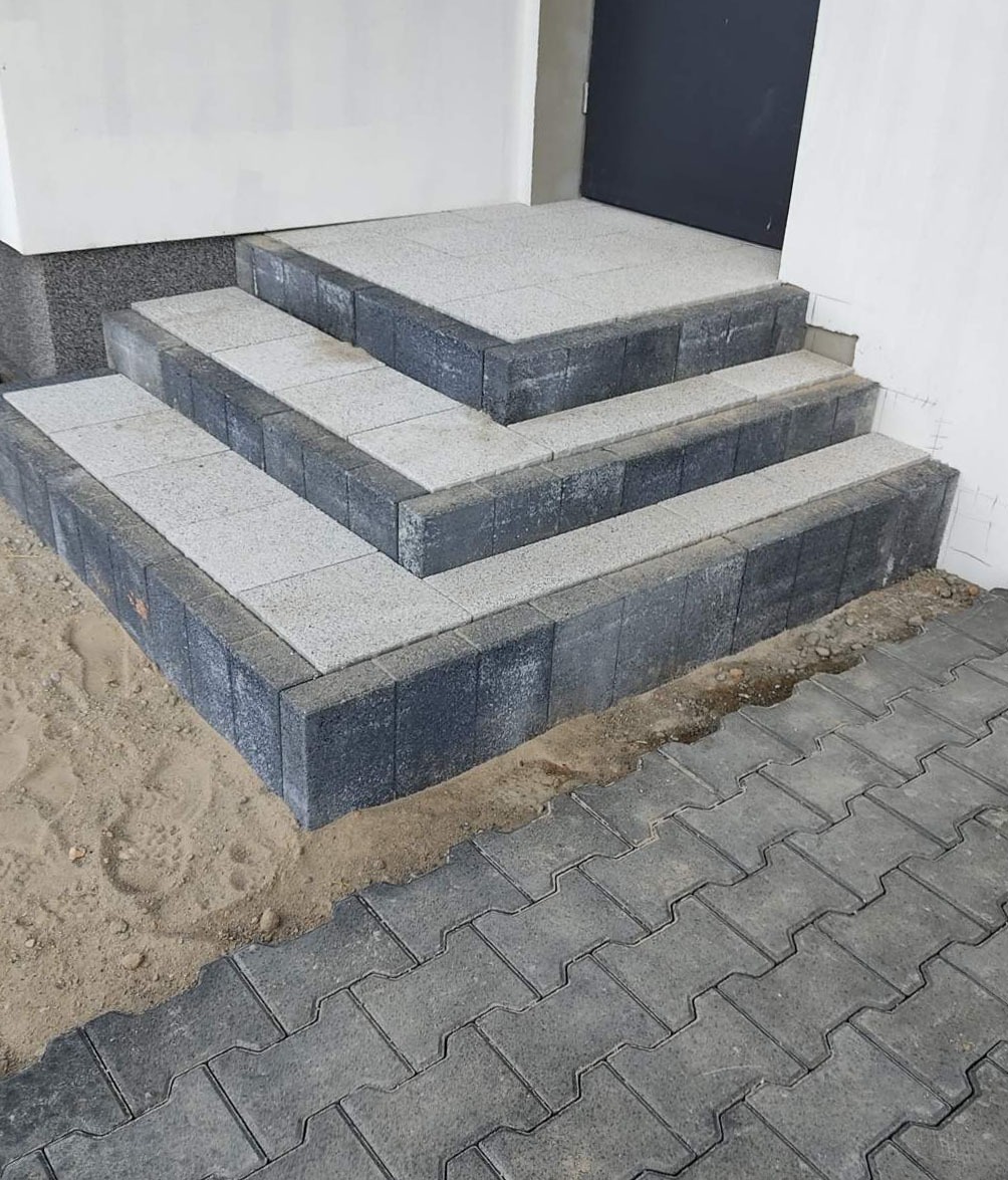 Schody wejściowe wyłożone szarą kostką brukową z ciemnoszarym obramowaniem, prowadzące do ciemnych drzwi, z widoczną kostką brukową na chodniku i piaskiem wokół.