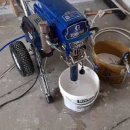 Agregat malarski Graco Texspray Mark VII z podłączonymi wężami i wiadrem farby Śnieżka, stojący na betonowej podłodze w trakcie przygotowań do malowania.