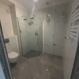 Wykończona łazienka z kabiną prysznicową o heksagonalnej podstawie, marmurowe płytki na ścianach i podłodze, podwieszana toaleta, grzejnik drabinkowy i nowoczesne oświetlenie.