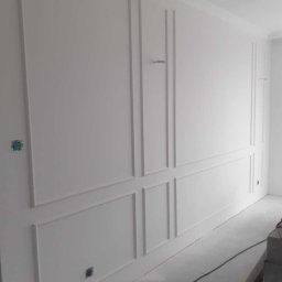 Prospray.pl - Biała ściana z dekoracyjnymi panelami ściennymi w stylu klasycznym, widoczne puszki elektryczne i kabel na podłodze, pomieszczenie w trakcie wykańczania.