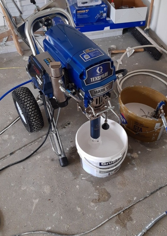 Agregat malarski Graco Texspray Mark VII z podłączonymi wężami i wiadrem farby Śnieżka, stojący na betonowej podłodze w trakcie przygotowań do malowania.