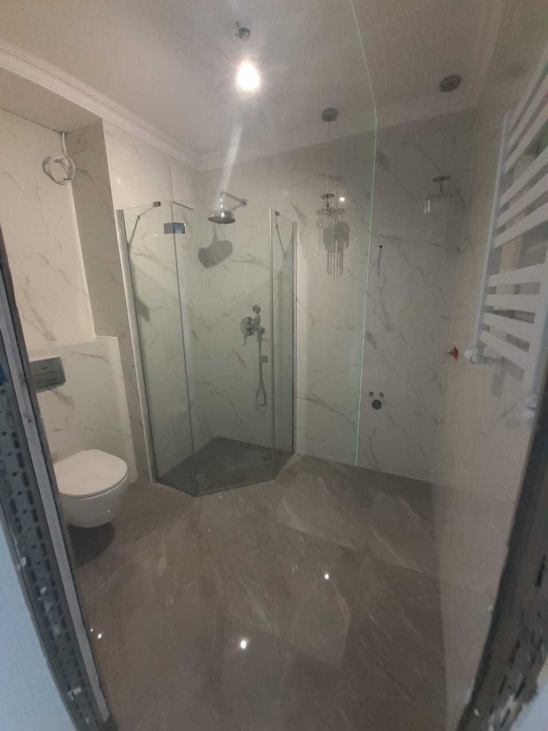 Wykończona łazienka z kabiną prysznicową o heksagonalnej podstawie, marmurowe płytki na ścianach i podłodze, podwieszana toaleta, grzejnik drabinkowy i nowoczesne oświetlenie.