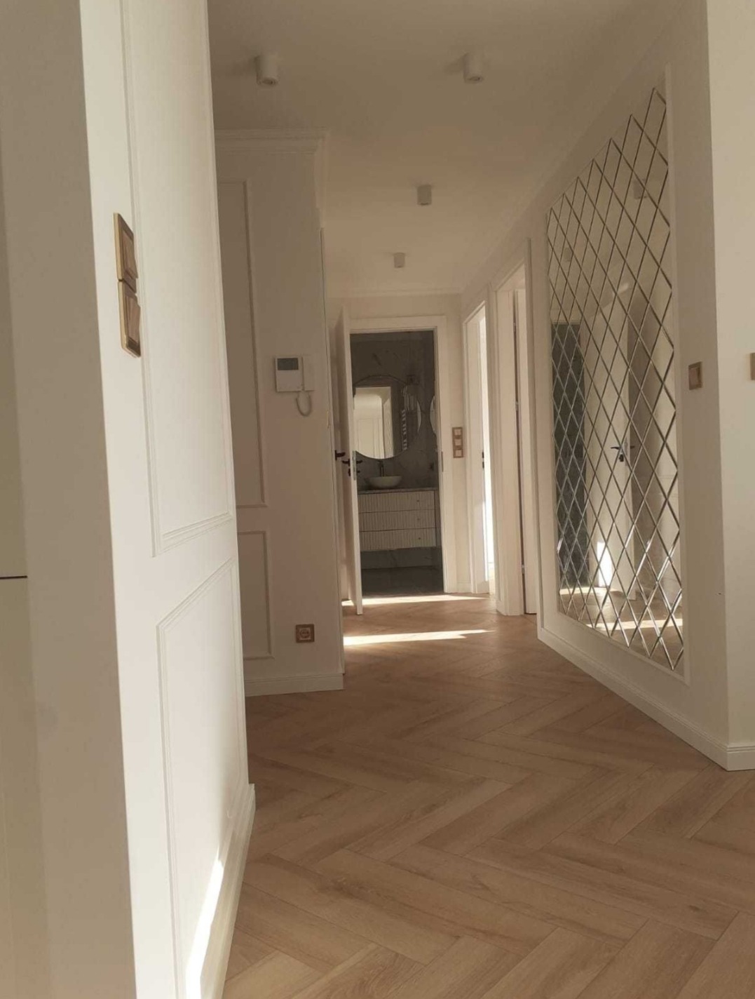 Jasny korytarz z parkietem w jodełkę, białe ściany z dekoracyjnymi panelami, lustro w romby i widok na łazienkę w tle.