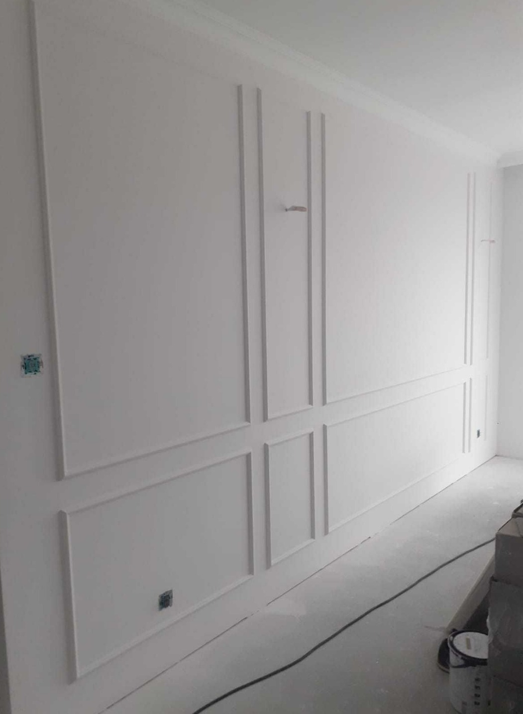 Biała ściana z dekoracyjnymi panelami ściennymi w stylu klasycznym, widoczne puszki elektryczne i kabel na podłodze, pomieszczenie w trakcie wykańczania.