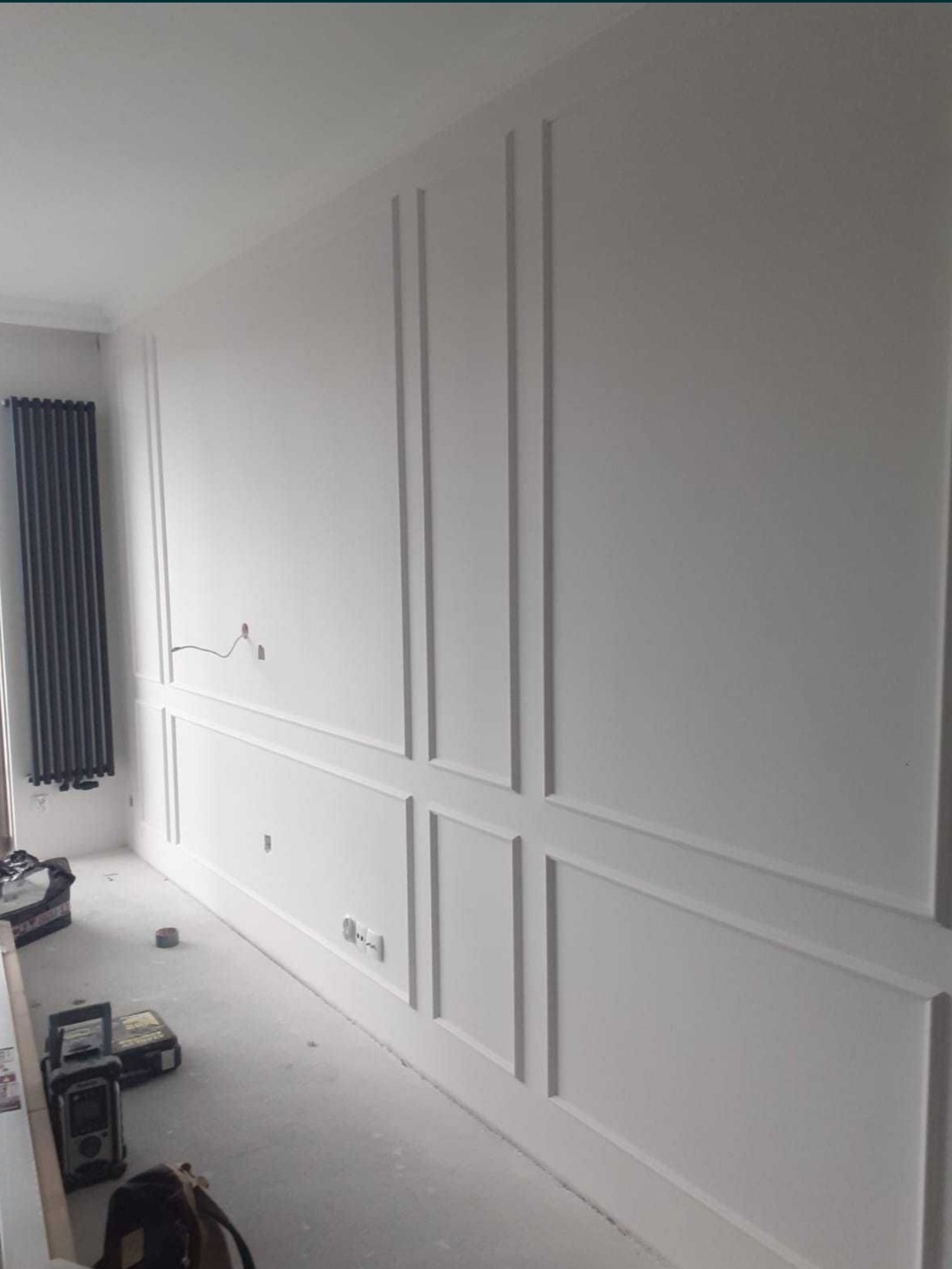 Ściana z dekoracyjnymi panelami ściennymi w trakcie montażu, widoczne gniazdka elektryczne, przewody i narzędzia na podłodze.