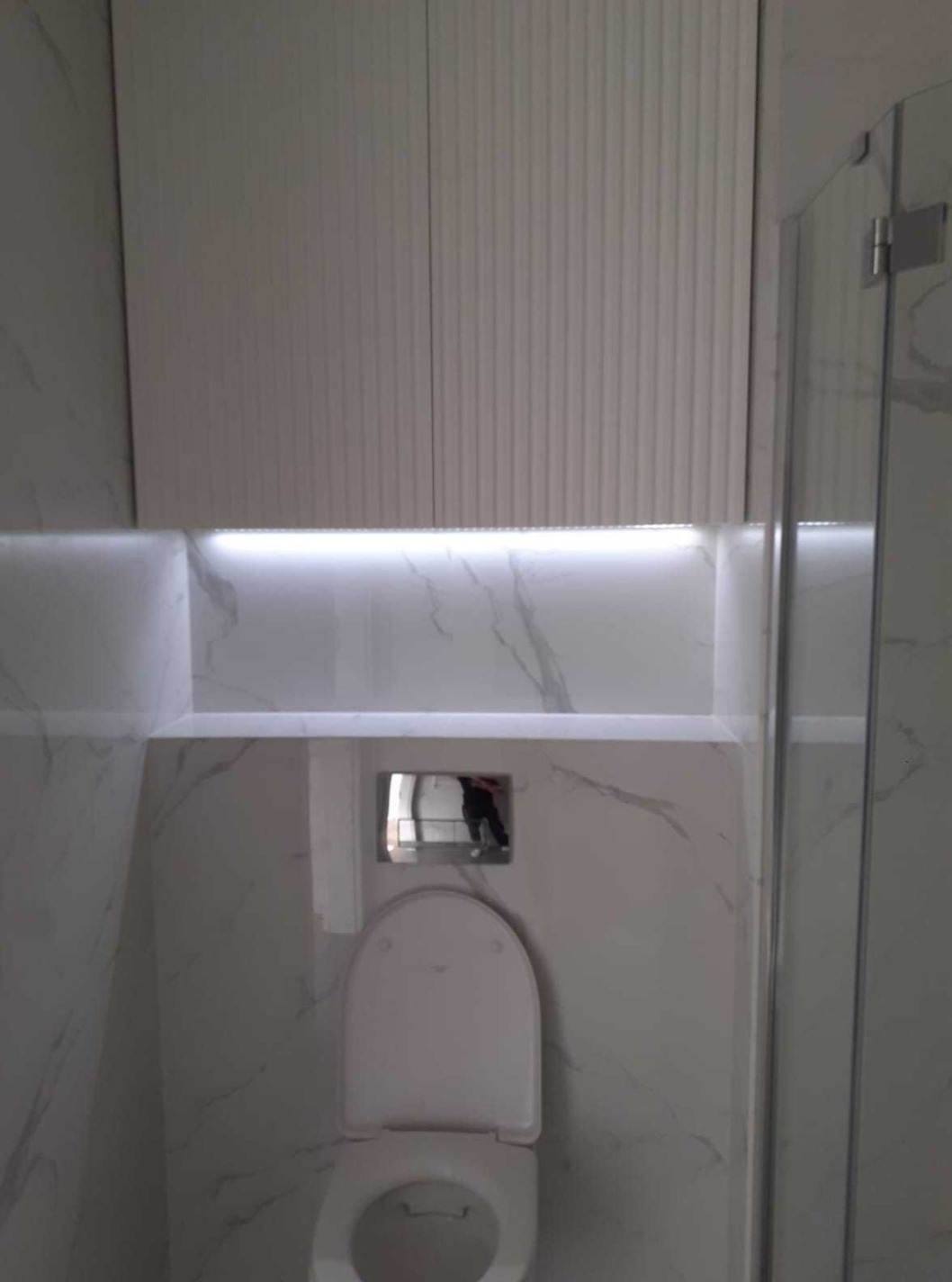 Wykończona łazienka z podświetlaną szafką nad toaletą, marmurowe płytki na ścianach i częściowo widoczna kabina prysznicowa z przezroczystego szkła.