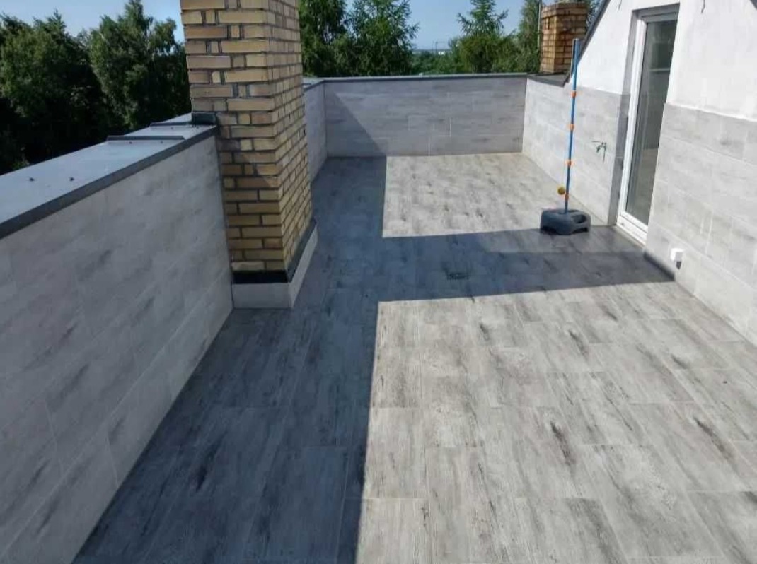 Wykończony taras na dachu z szarymi płytkami imitującymi drewno, ceglany komin i szare ściany, widok na zewnątrz.