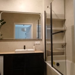 VIKTOR TALAILO Twój Dom Glazurnictwo - Nowoczesna łazienka po remoncie: czarna szafka z drewnianym blatem, umywalka, lustro z zaokrąglonymi rogami i szklany parawan nawannowy z czarnymi ramami.