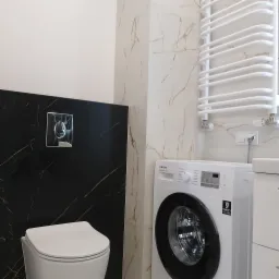 Nowoczesna łazienka z czarnymi i białymi płytkami imitującymi marmur, wisząca toaleta, grzejnik drabinkowy i pralka Samsung. Minimalistyczny design.
