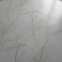 Ułożone, jasne płytki podłogowe z marmurkowym wzorem, widoczne fugi i fragment okna.