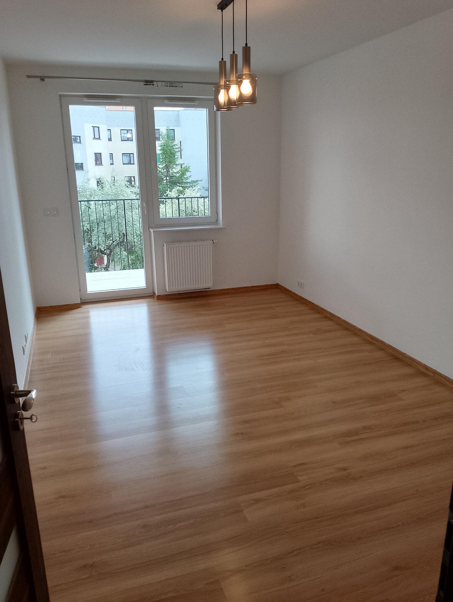 Pusty, jasny pokój z panelami podłogowymi w odcieniach brązu, widok na balkon z zielenią i nowoczesną lampą wiszącą z trzema szklanymi kloszami.