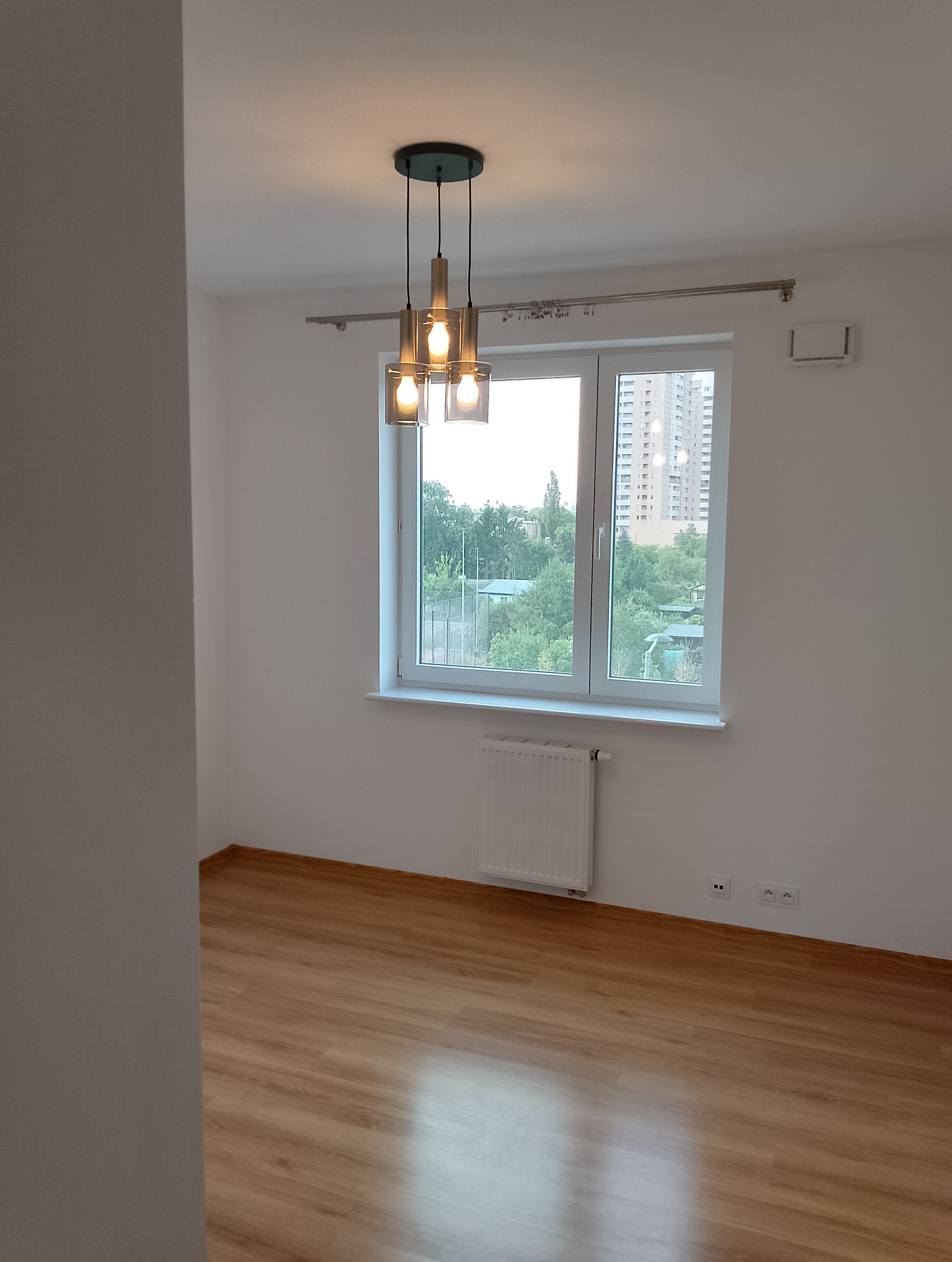 Pusty pokój z drewnianą podłogą, białymi ścianami, grzejnikiem pod oknem z widokiem na zieleń i budynki, oraz nowoczesną lampą wiszącą z trzema kloszami.