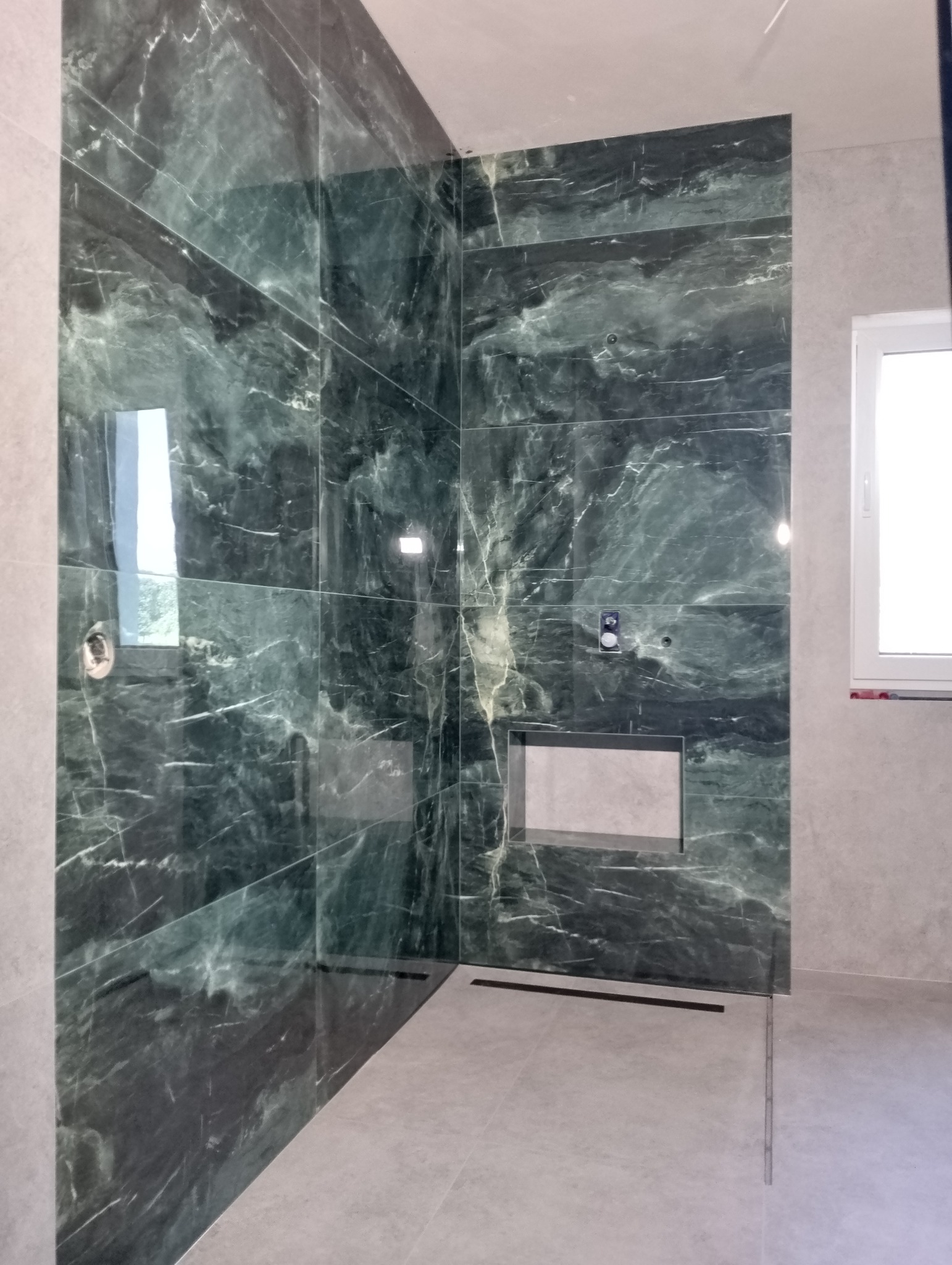 Wykończona kabina prysznicowa z zielonymi płytkami imitującymi marmur, z odpływem liniowym w podłodze i wnęką w ścianie wyłożoną jasnymi płytkami.