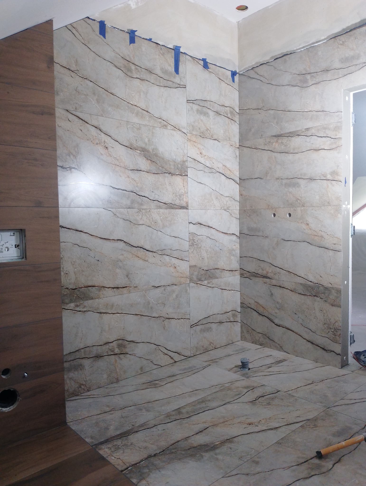 Łazienka w trakcie układania płytek: beżowe płytki imitujące marmur z ciemnymi żyłkami na ścianach i podłodze, zabezpieczone niebieską taśmą malarską przy suficie, widoczny odpływ liniowy...