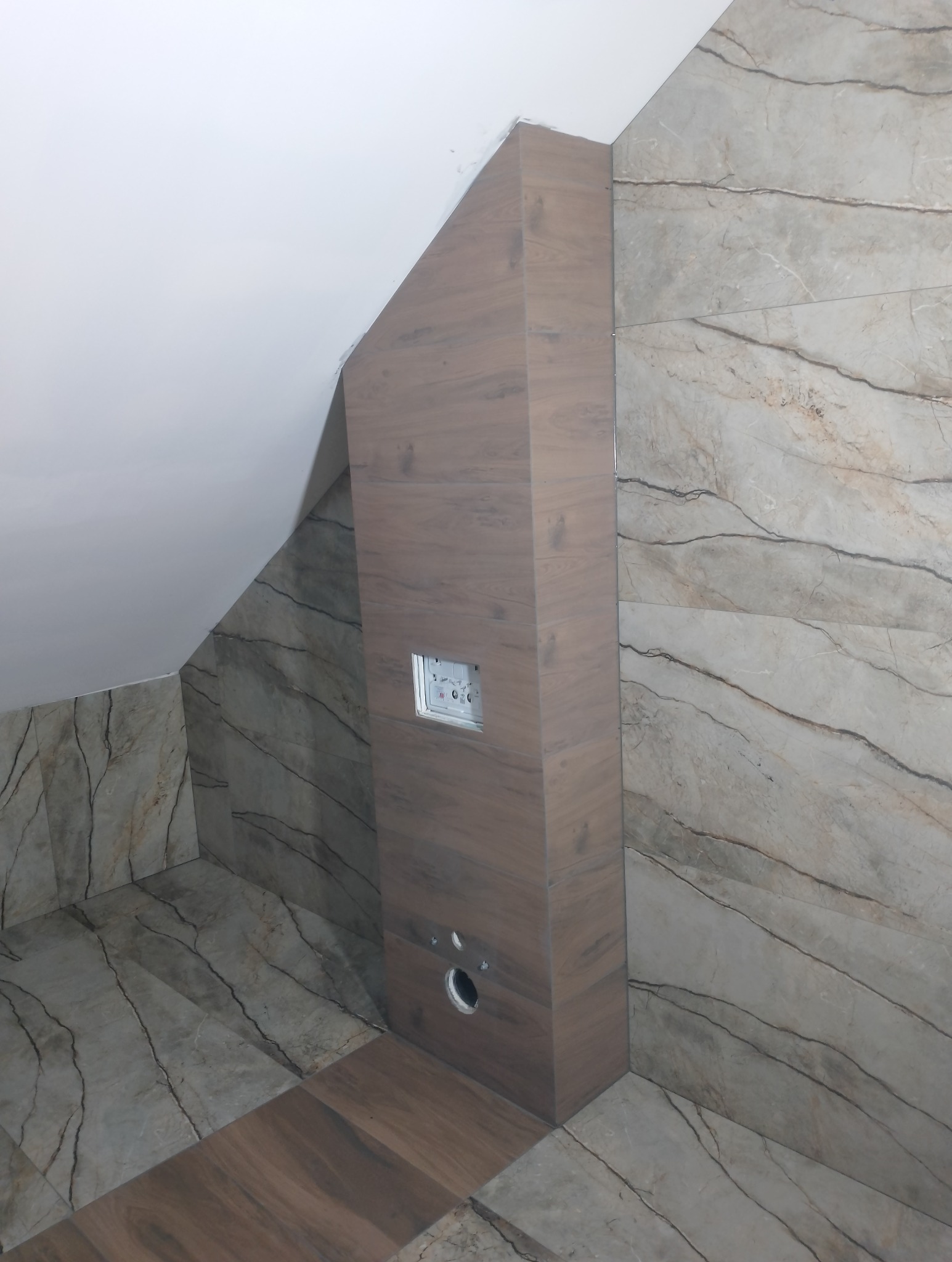 Wykończona łazienka na poddaszu ze skośnym sufitem, marmurowe płytki na ścianach i podłodze, zabudowa geberitu wykończona płytkami imitującymi drewno.