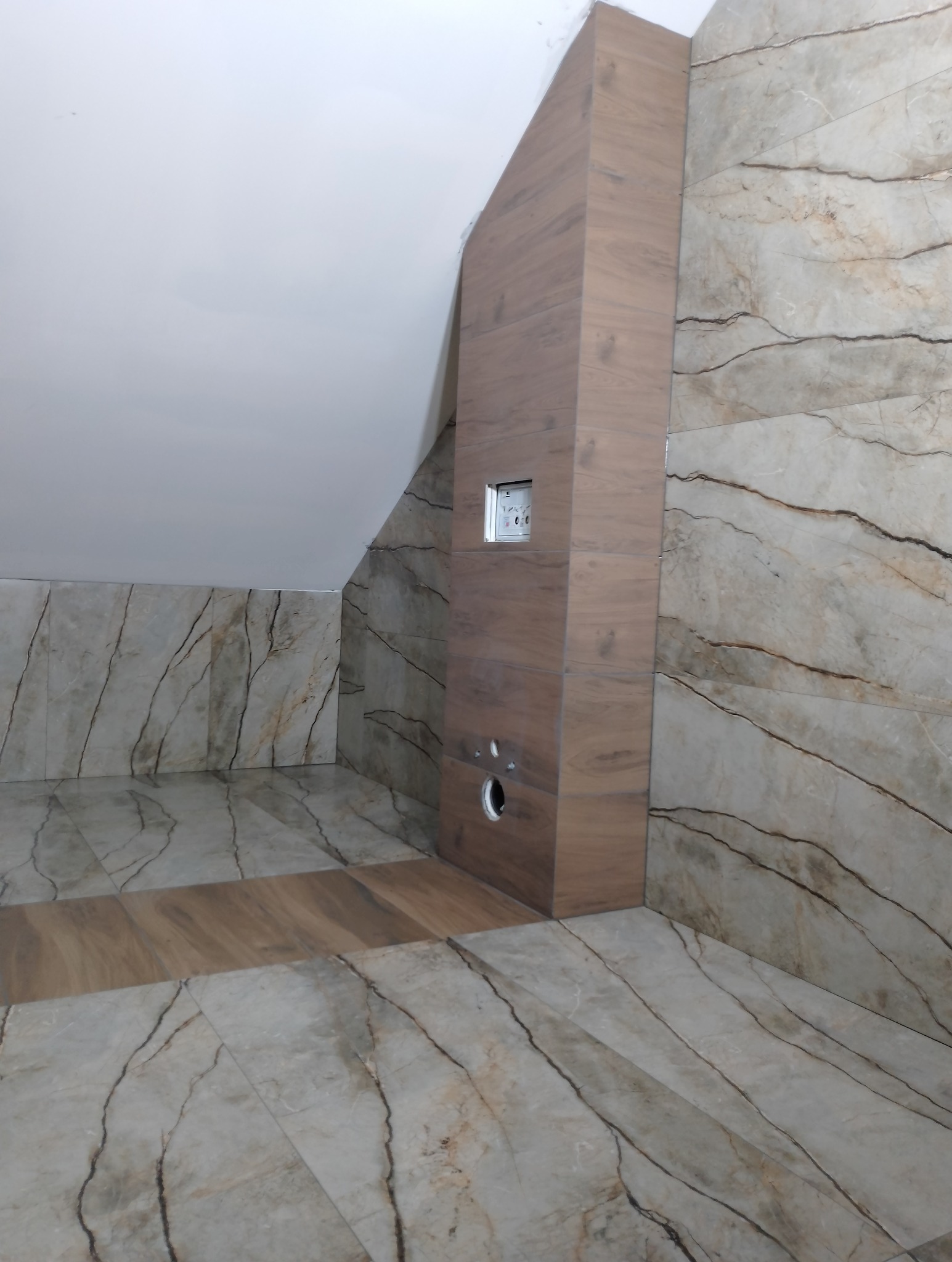 Wykończona łazienka na poddaszu ze skośnym sufitem, marmurowe płytki na ścianach i podłodze, zabudowa z drewnopodobnych płytek maskująca instalację sanitarną z otworem rewizyjnym i przyciskiem...