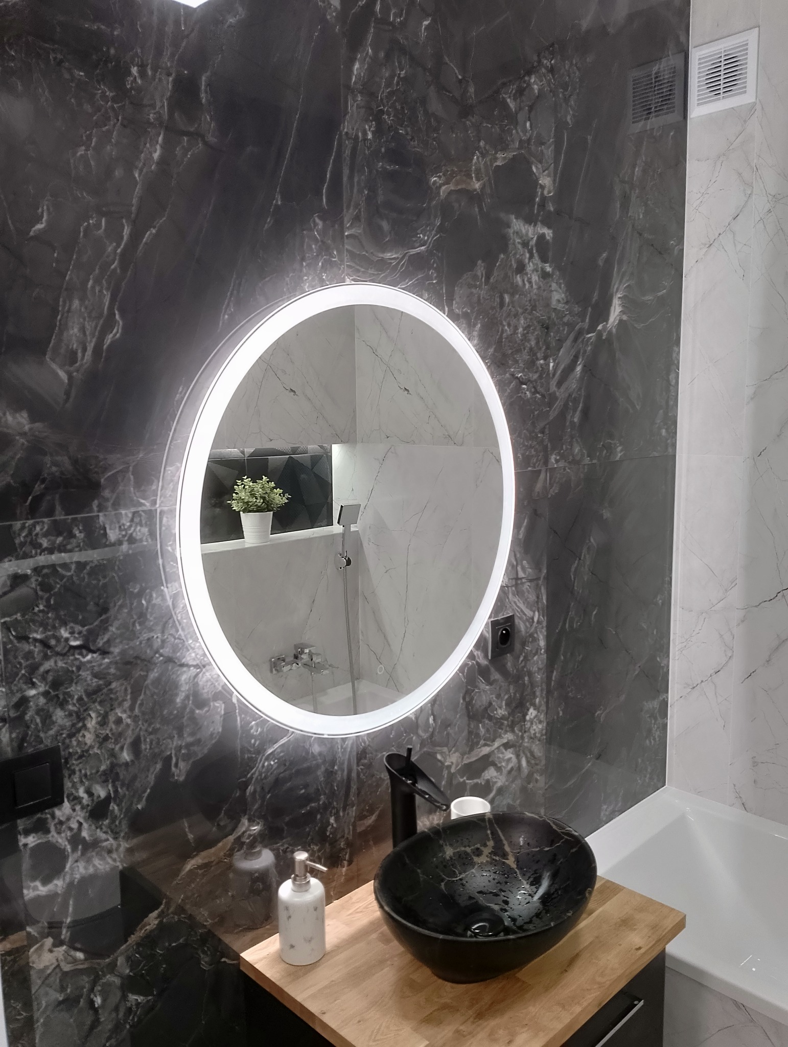 Nowoczesna łazienka z czarnymi marmurowymi płytkami, okrągłym lustrem z podświetleniem LED, czarną umywalką na drewnianym blacie i białą wanną.