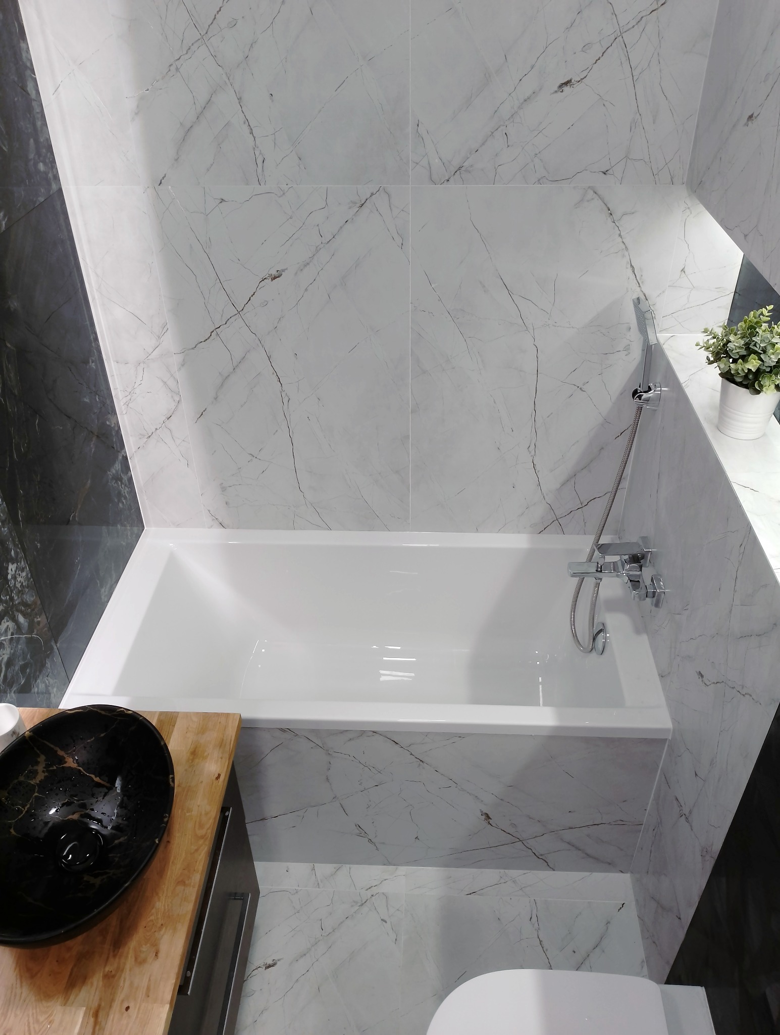 Nowoczesna łazienka z białą wanną obudowaną marmurowymi płytkami, czarną umywalką na drewnianym blacie i minimalistyczną armaturą.