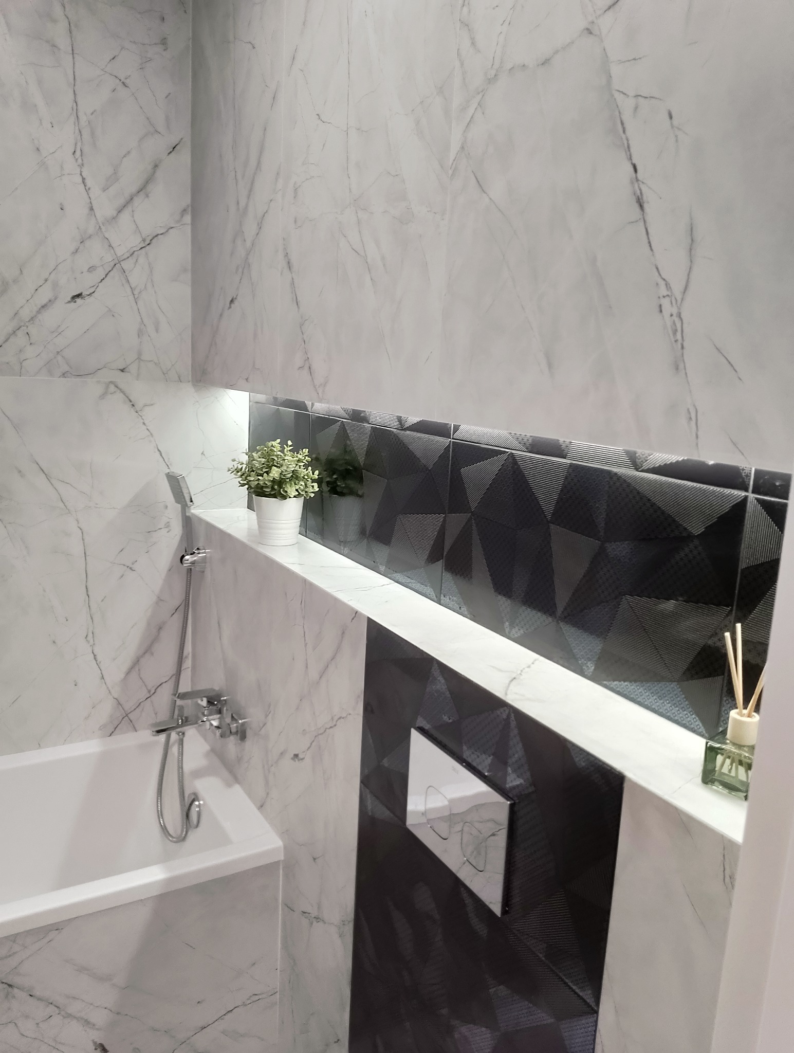 Nowoczesna łazienka z wanną, baterią prysznicową, marmurowymi płytkami na ścianach, czarnymi płytkami z geometrycznym wzorem oraz półką z oświetleniem LED i dekoracjami.