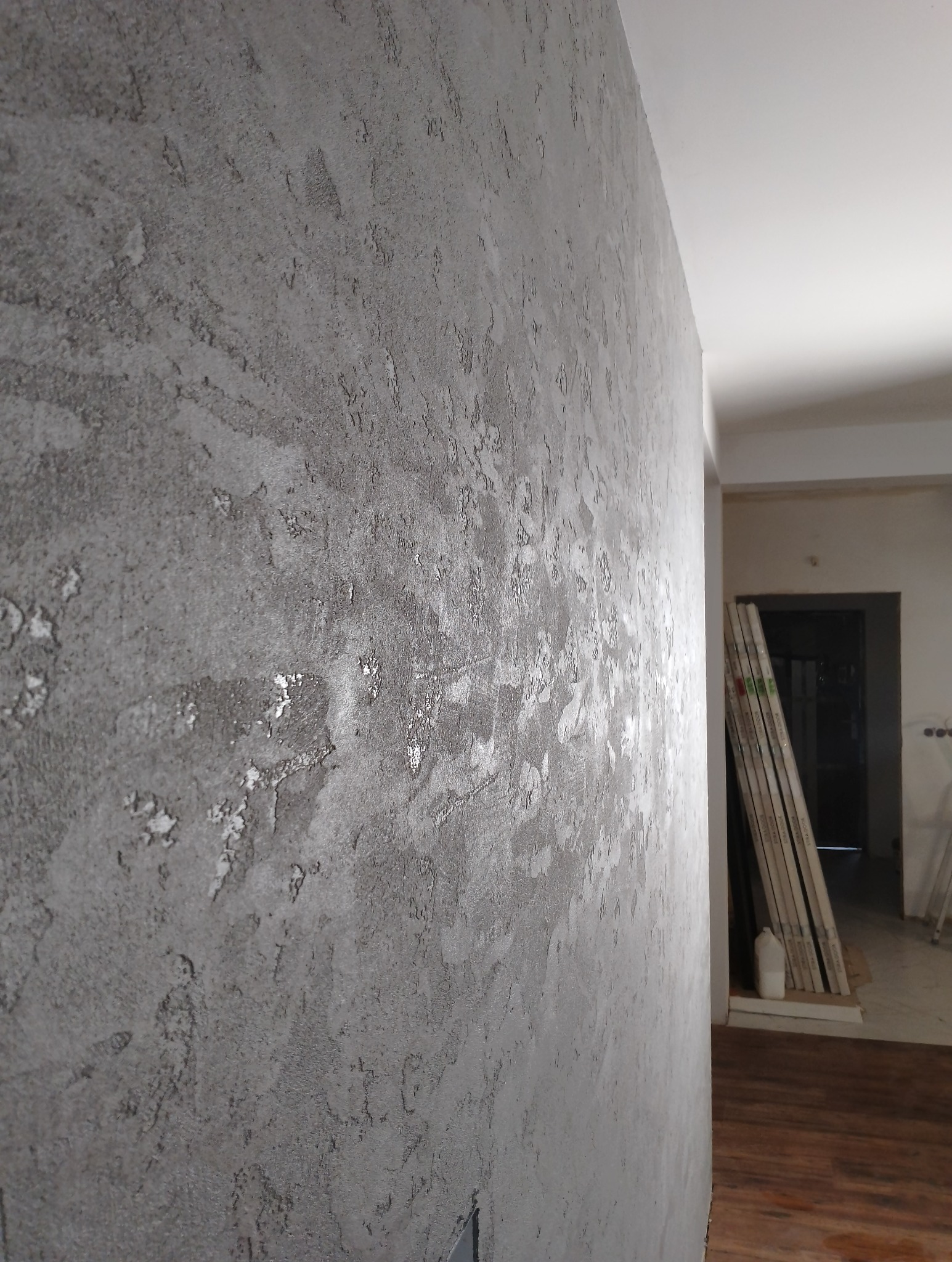 Szara ściana z dekoracyjną fakturą w stylu betonowym, widok z boku, w tle podłoga z paneli i materiały budowlane przy futrynie drzwi.