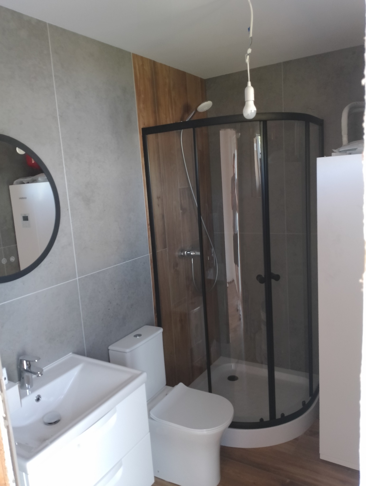 Wykończona łazienka w Płońsku: szare płytki, kabina prysznicowa z czarnymi ramami, biała umywalka i toaleta. Nowoczesny design.