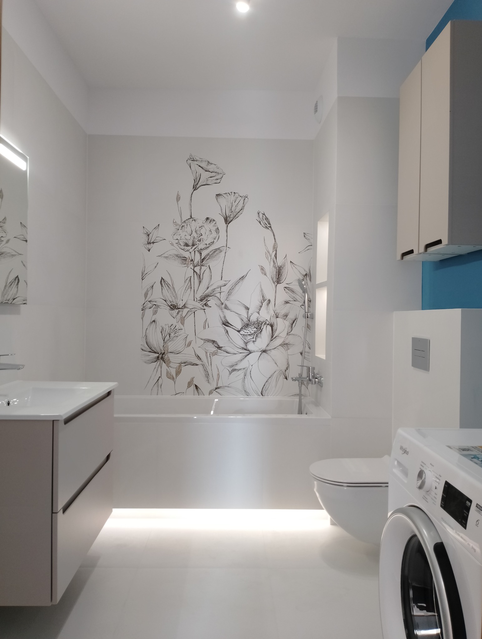Jasna łazienka z akcentem kwiatowym na ścianie, podświetleniem pod wanną i szafką umywalkową. Minimalistyczny design z pralką w rogu.