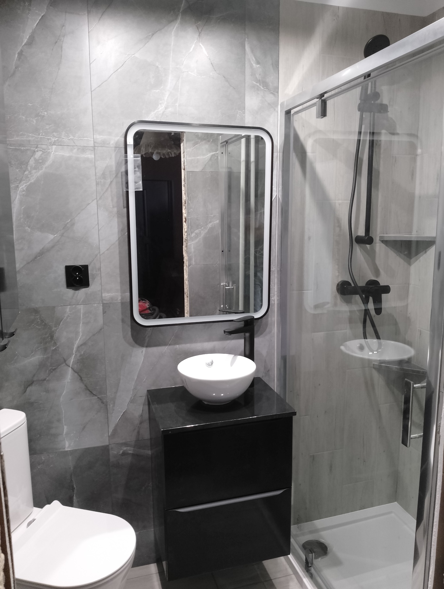 Nowoczesna łazienka z szarymi płytkami imitującymi marmur, czarną armaturą i kabiną prysznicową. Umywalka nablatowa na szafce. W lustrze odbicie wnętrza.